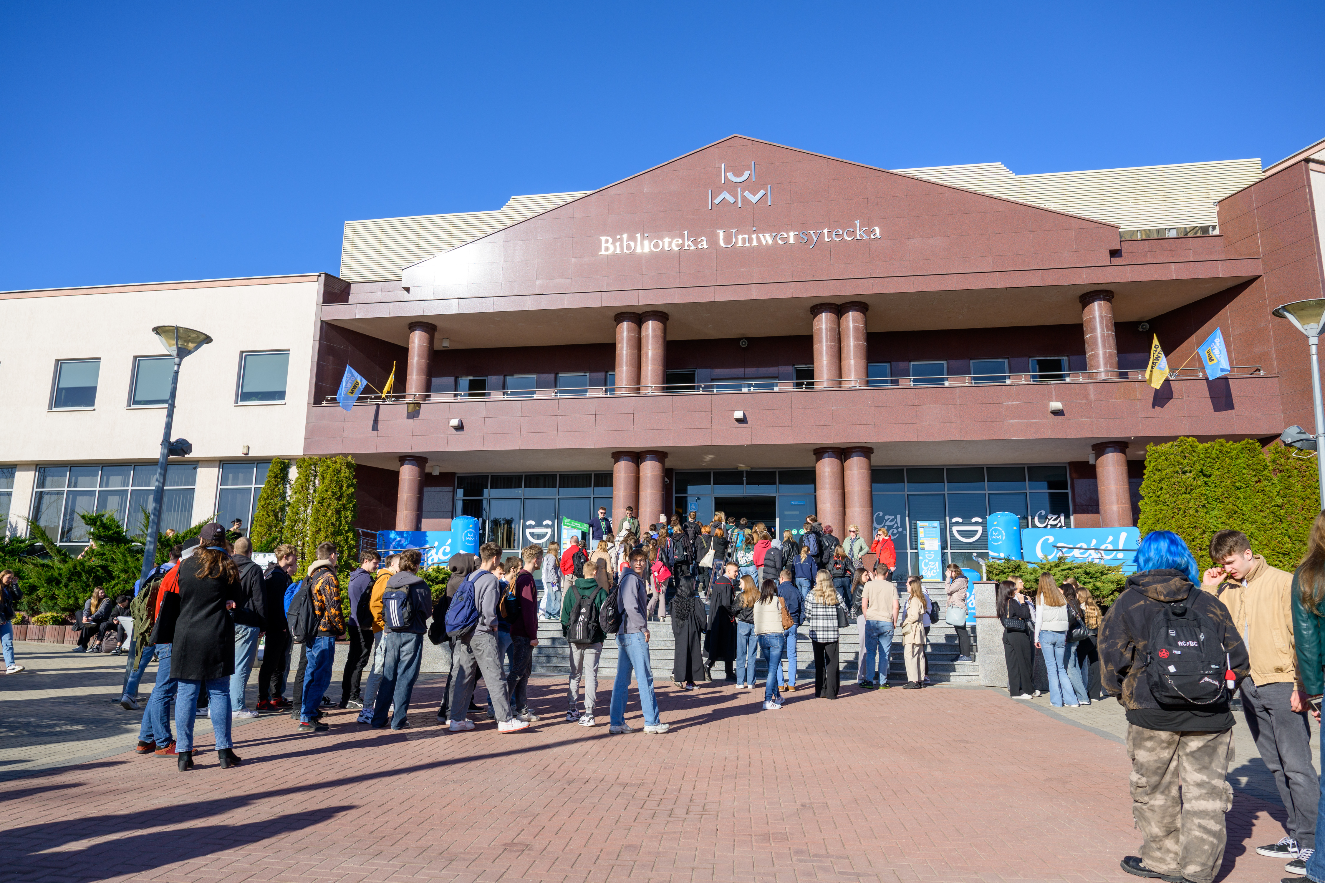 Uczestnicy Dnia Otwartego UWM gromadzą się przed wejściem do Biblioteki Uniwersyteckiej. Na schodach i placu przed budynkiem stoi grupa młodych osób, a nad wejściem widoczny jest napis „Biblioteka Uniwersytecka”. Zdjęcie wykonano w słoneczny dzień.