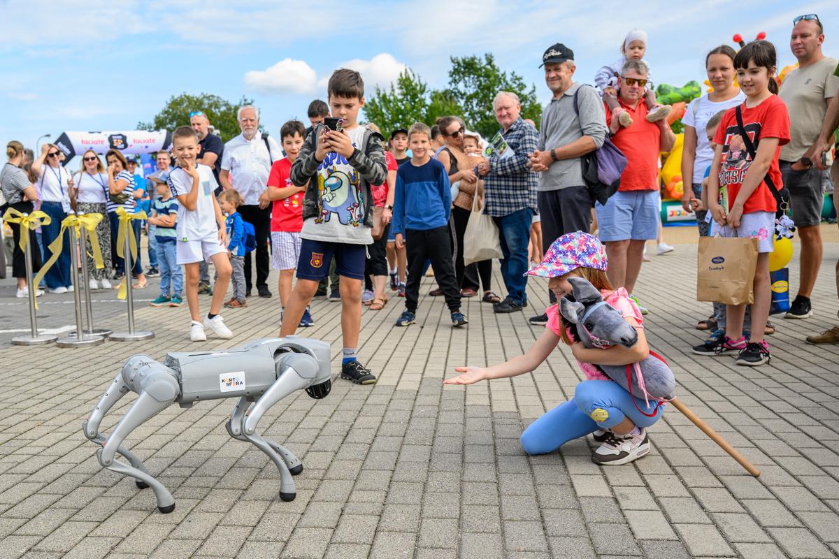 Zdjęcie przedstawia dziecko i robota-psa. W tle stoją inne osoby