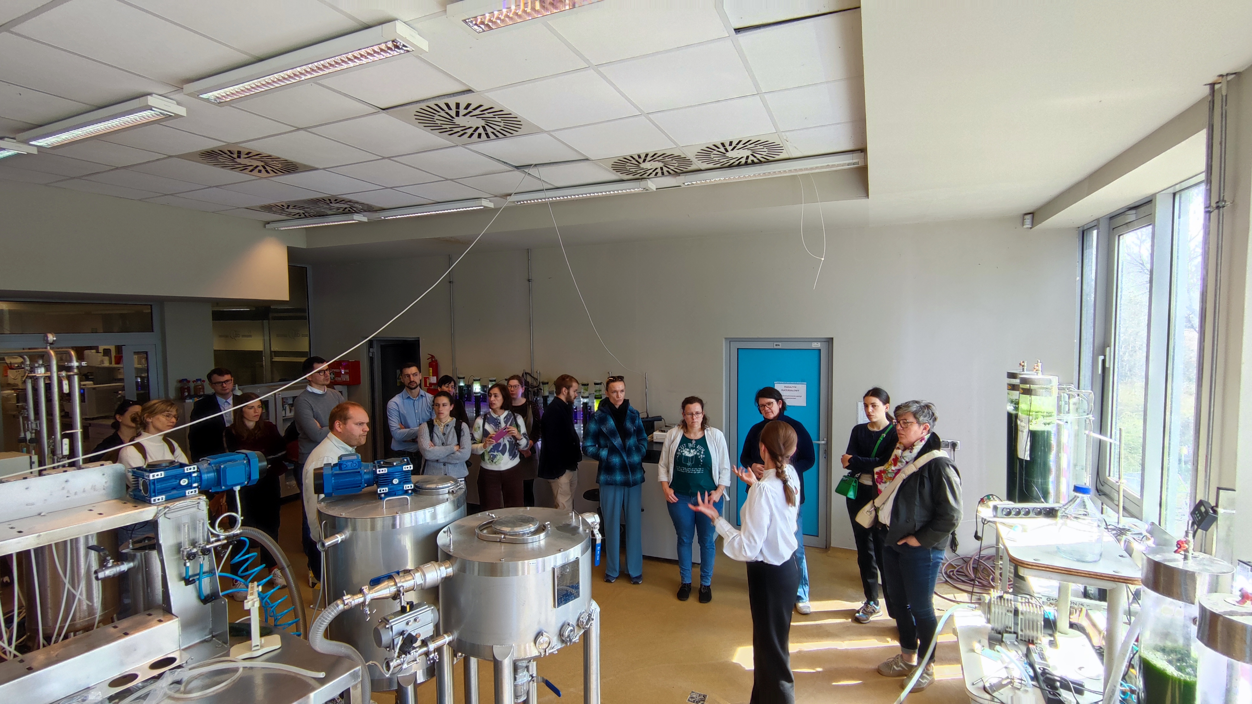 Uczestnicy zgromadzeni w laboratorium Uniwersytetu Warmińsko-Mazurskiego, słuchający osoby oprowadzającej, która objaśnia aparaturę inżynierii środowiska wykorzystywaną do uzdatniania wody i badań.