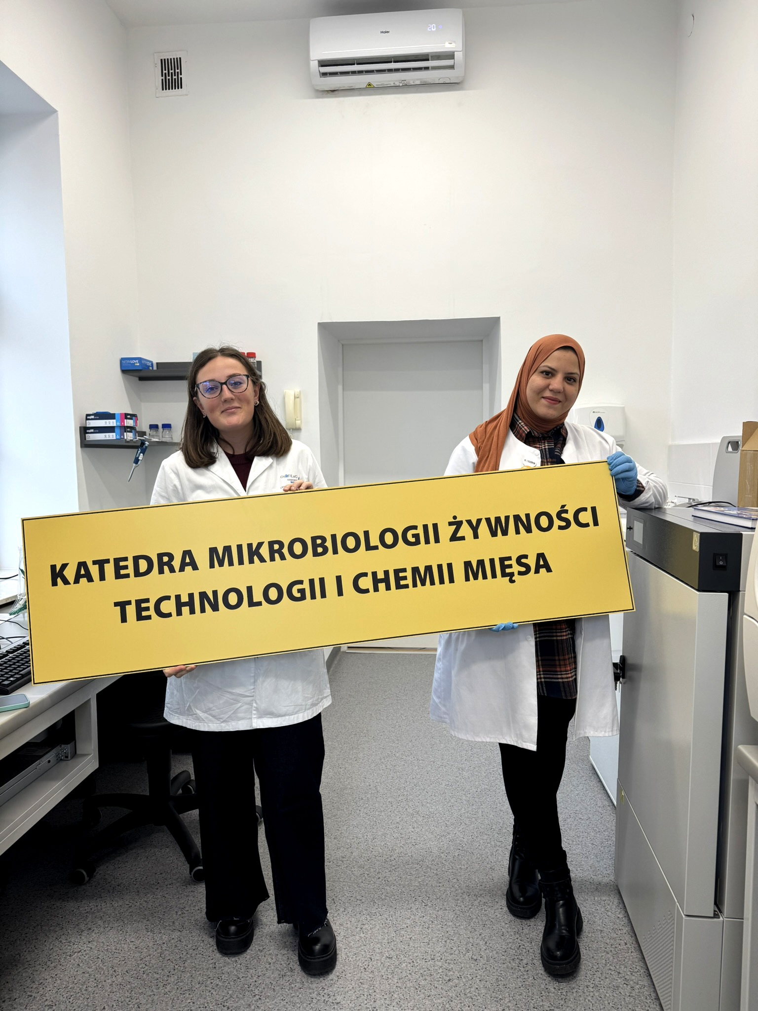 doktorantki stoją w laboratorium i trzymają tablicę z napisem Katedra Mikrobiologii Żywności, Technologii i Chemii Mięsa