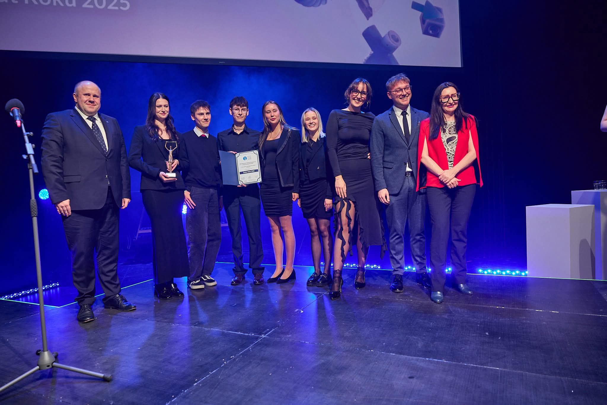 osoby stoją na scenie, trzymają dyplom i statuetkę