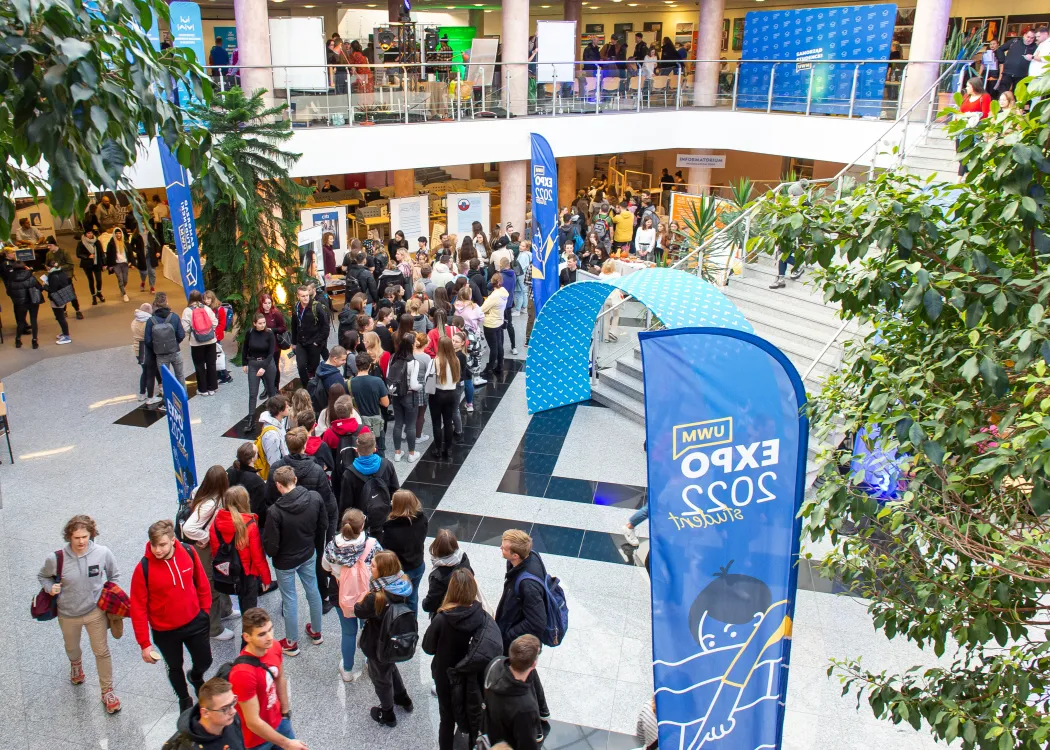 widok z góry na studentów stojących w bibliotece uniwersyteckiej podczas wydarzenia Student Expo, po prawej stoją niebieski flagi