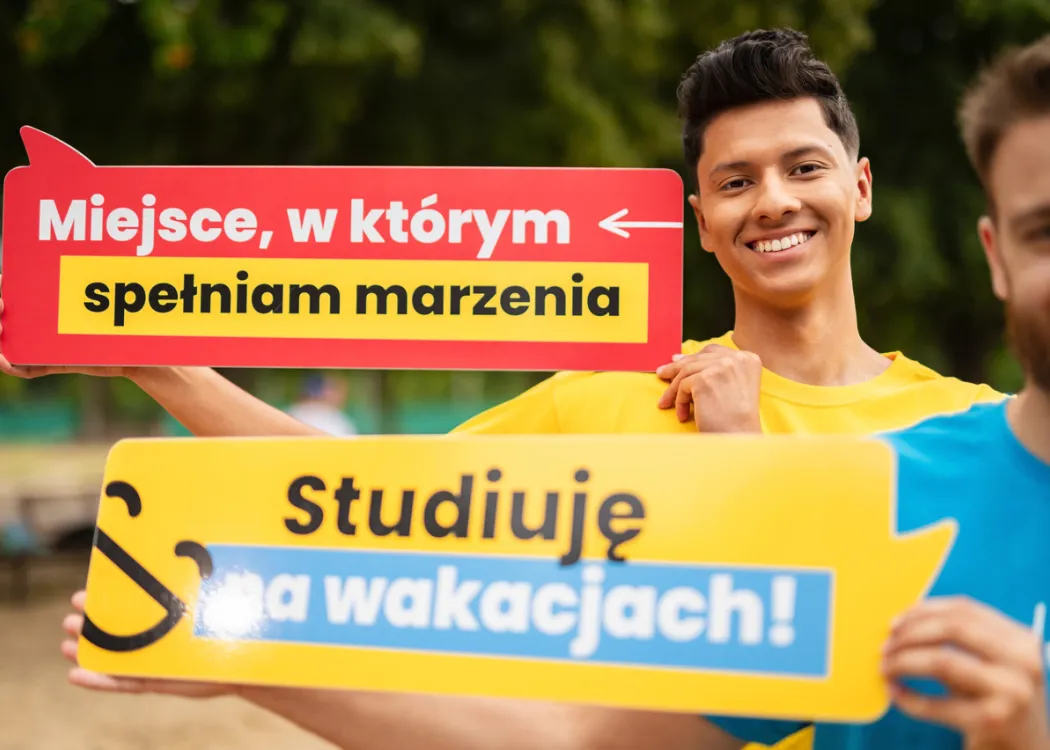 Dwaj młodzi mężczyźni pozują z tabliczkami: Miejsce, w którym spełniam marzenia i Studiuję na wakacjach!