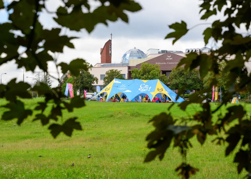 namiot w kolorach niebiesko-żółtych, z napisem cześć i uśmiechniętą buźką, zielona trawa, w tle biblioteka uwm