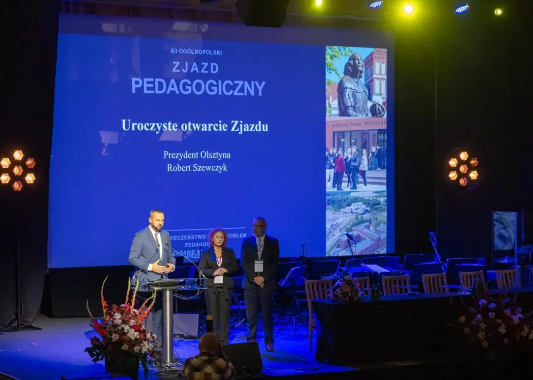 Zjazd pedagogiczny: trzy osoby stoją na scenie, w tle niebieski ekran