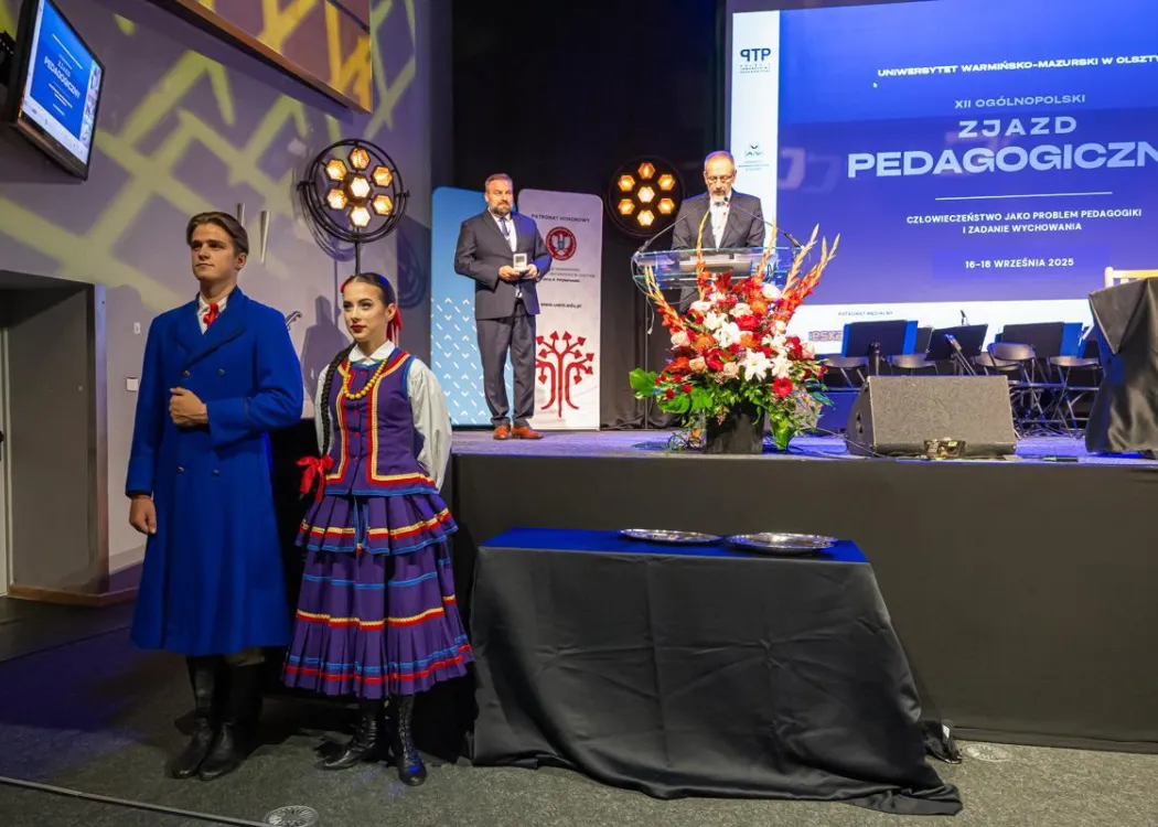 Zjazd pedagogiczny: para w strojach ludowych obok sceny, na scenie dwóch meżczyzn