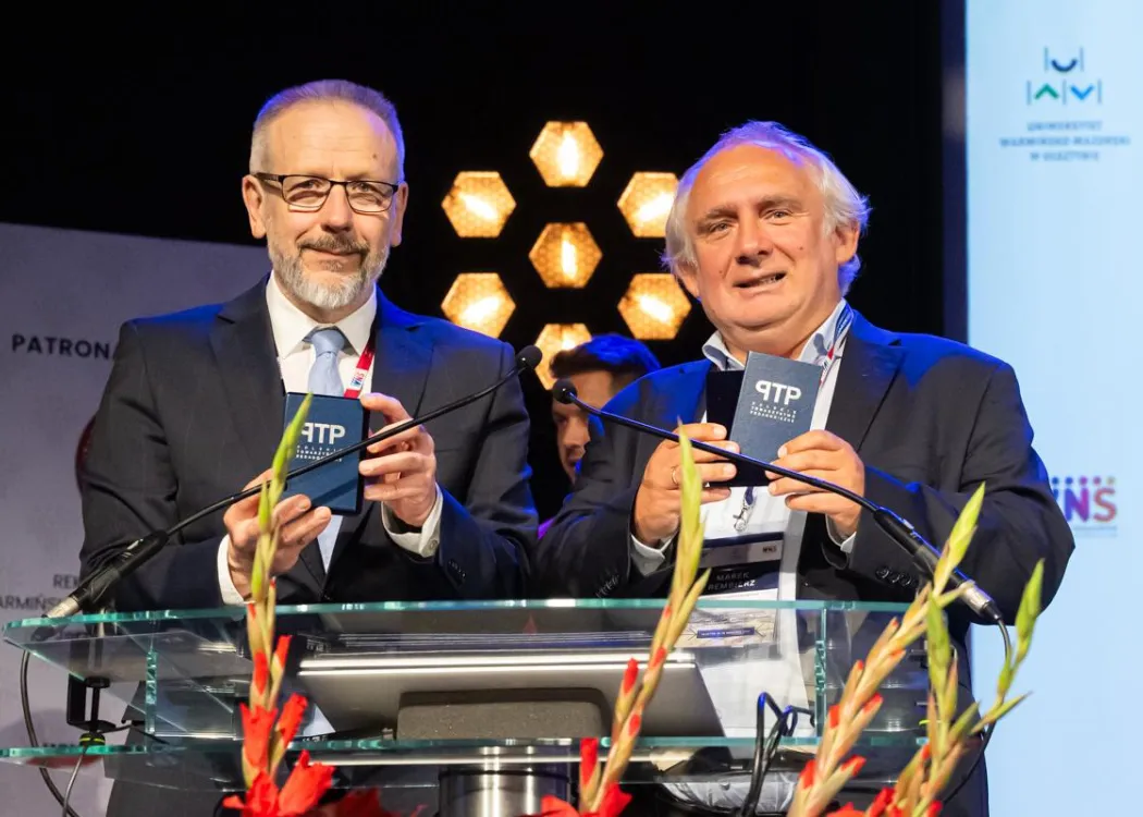 Zjazd pedagogiczny: dwóch mężczyzn pokazuje przy mównicy odznaczenia