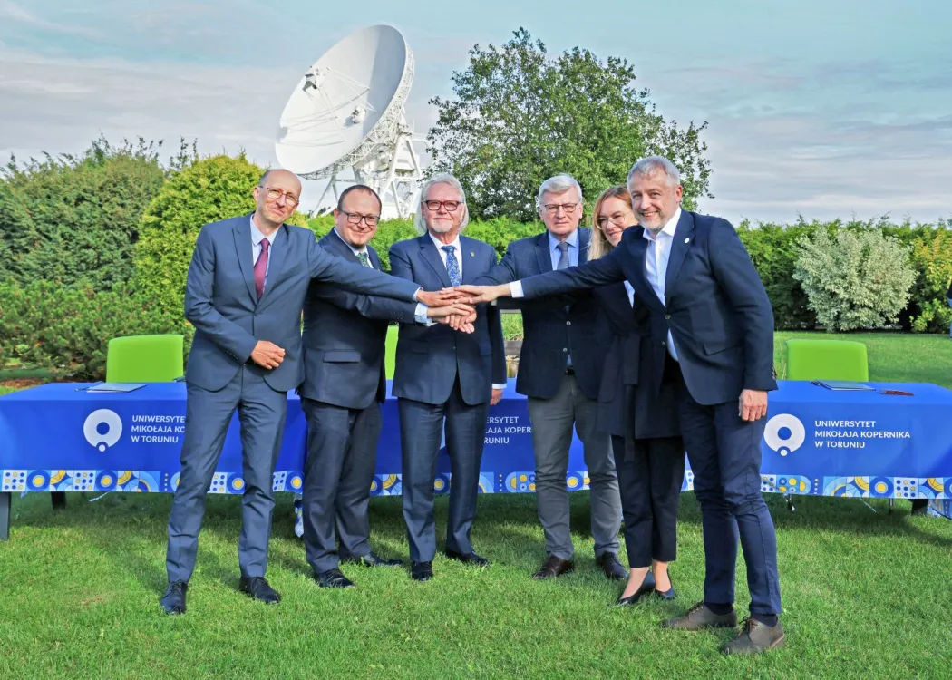 Sześcioro przedstawicieli polskiego konsorcjum projektu SKA Observatory stoi na tle anteny i trzyma dłonie położone jedna na drugiej