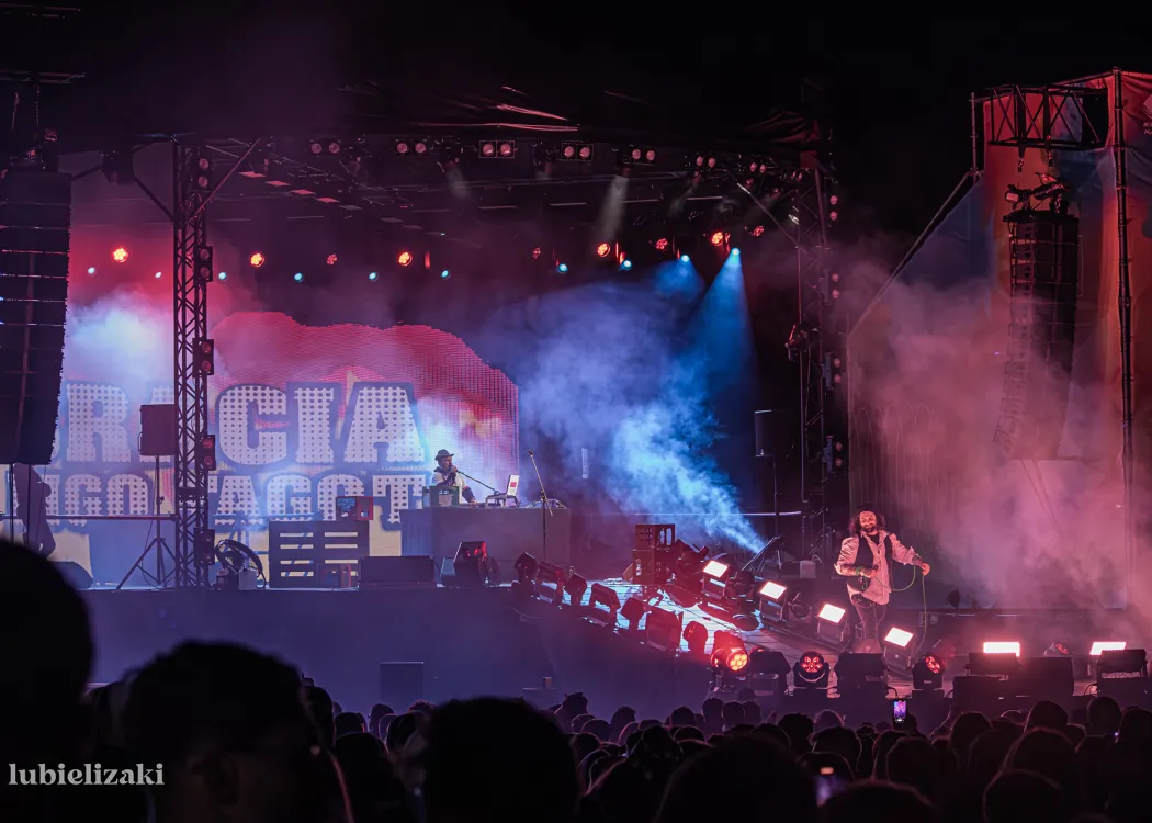 Scena z kolorowym oświetleniem na której występuje dwóch artystów - jeden na przodzie sceny śpiewa do mikrofonu, drugi z tyłu sceny gra na keyboardzie