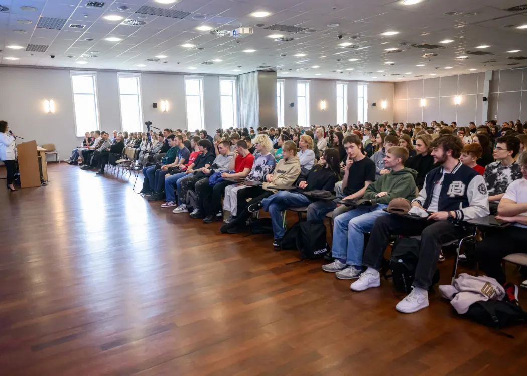 Widok na dużą salę wykładową wypełnioną publicznością siedzącą w rzędach krzeseł; na pierwszym planie prowadząca stojąca przy mównicy i przemawiająca do uczestników.