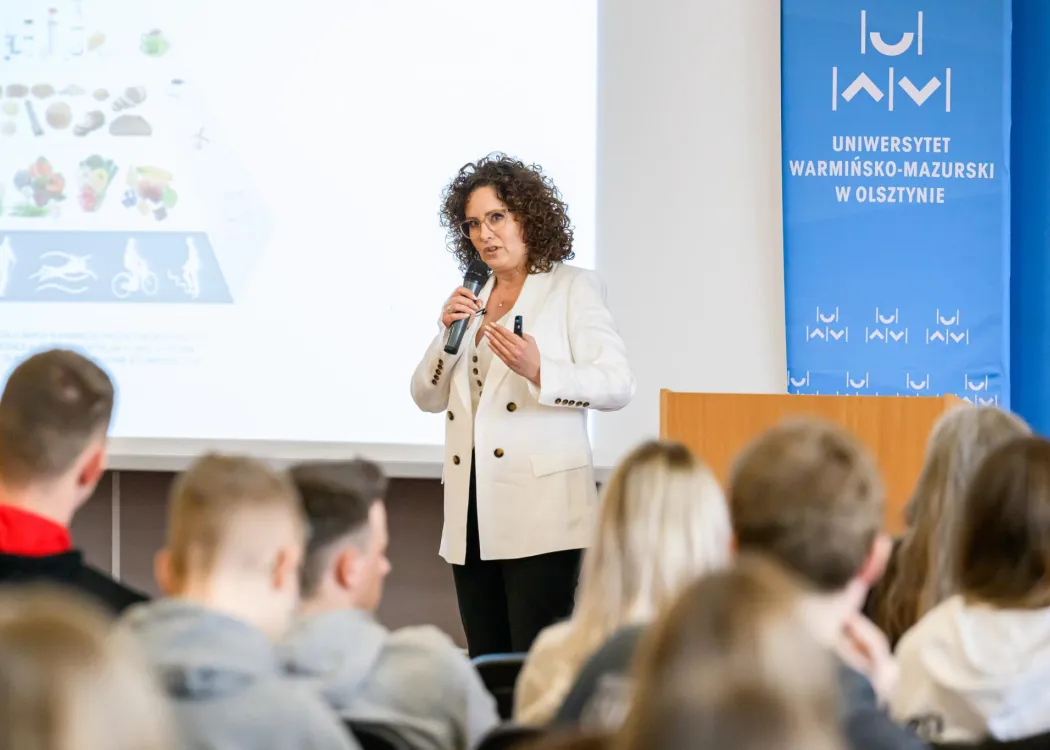 Prelegentka z mikrofonem przemawia do zgromadzonej publiczności; w tle ekran z prezentacją oraz baner Uniwersytetu Warmińsko-Mazurskiego w Olsztynie.