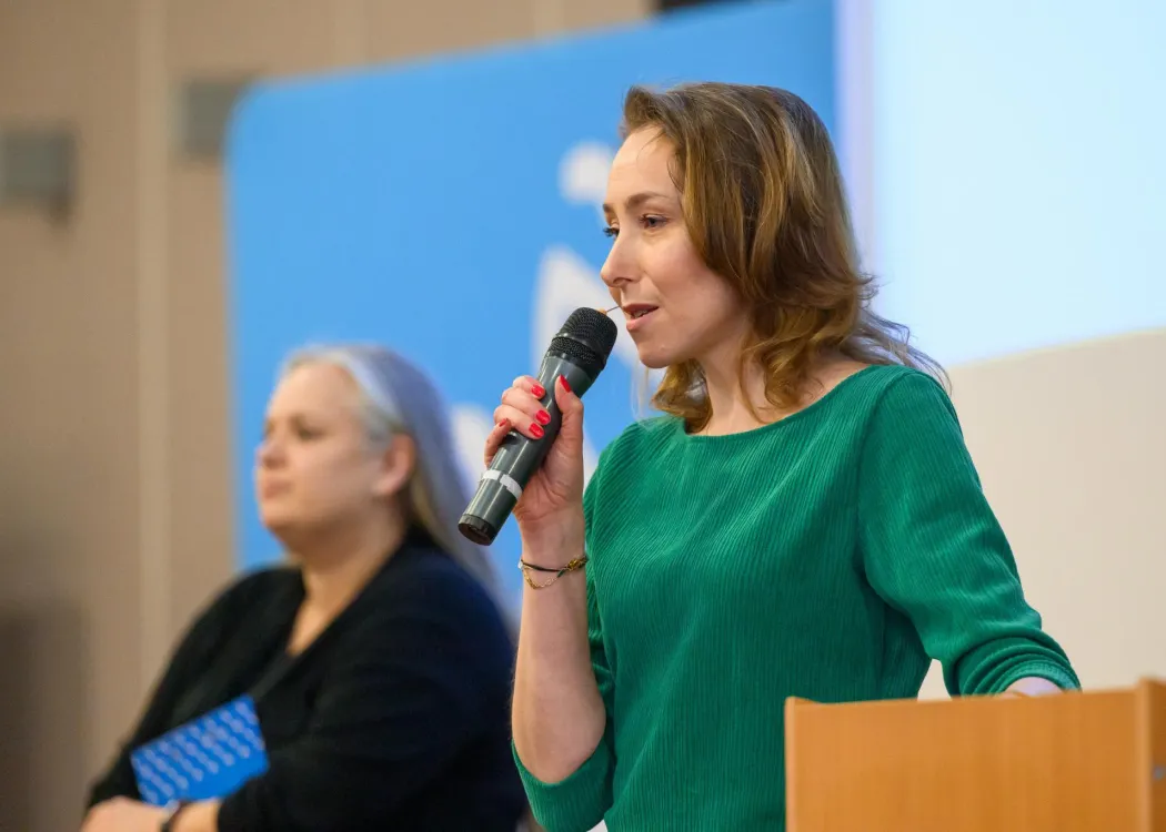 Kobieta stojąca przy mównicy trzyma mikrofon i przemawia do uczestników wydarzenia; w tle druga osoba siedząca przy stole oraz ekran prezentacyjny.