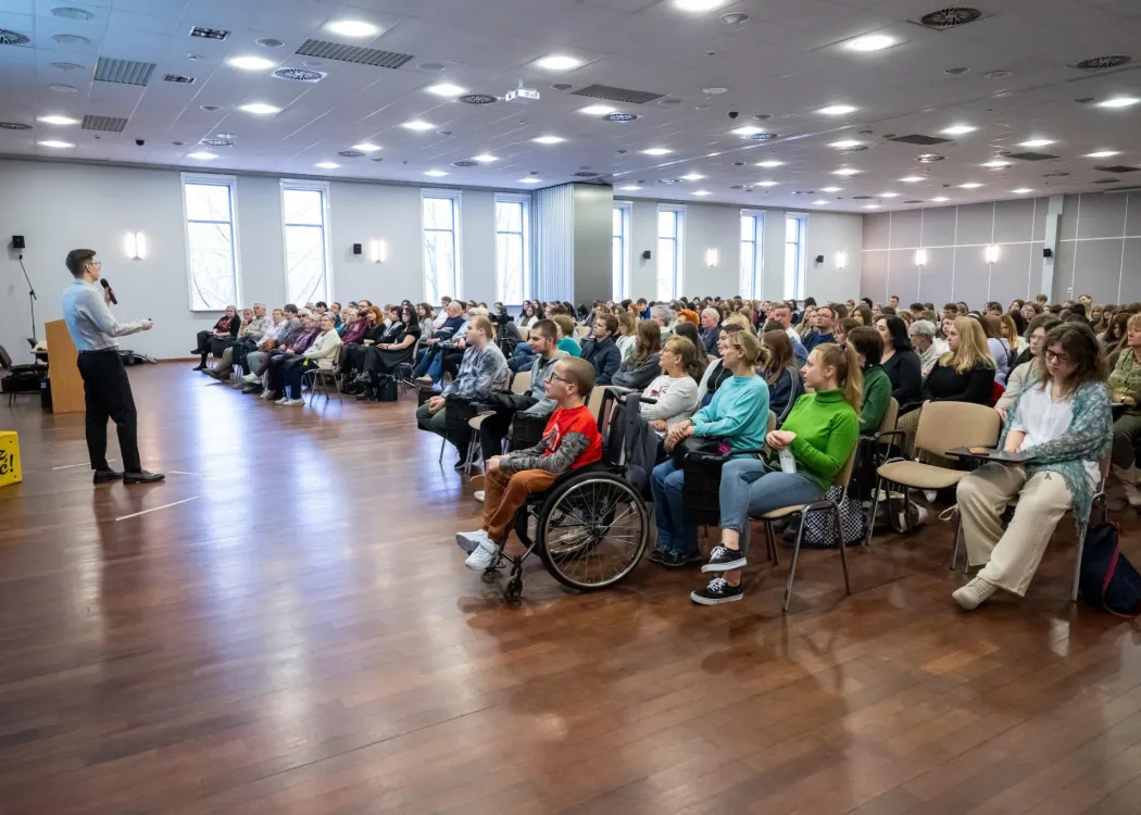 Szeroki plan sali wykładowej z liczną publicznością, w tym osobą poruszającą się na wózku, słuchającą prelegenta stojącego z mikrofonem.
