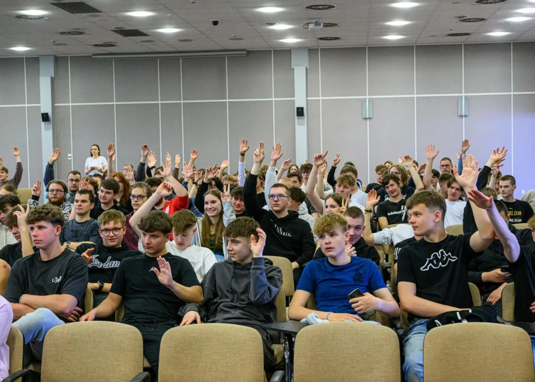 Grupa młodych uczestników siedzących na widowni podnosi ręce, zgłaszając się do odpowiedzi lub udziału w dyskusji.