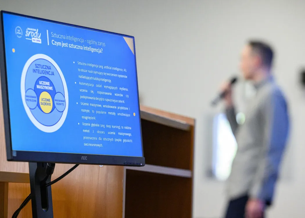 Ekran z prezentacją na temat sztucznej inteligencji i uczenia maszynowego; w tle rozmyta sylwetka prelegenta mówiącego do mikrofonu.