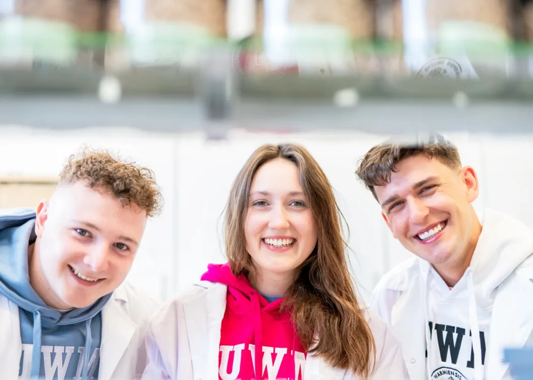 Troje młodych osób w bluzach UWM i białych fartuchach pozuje w laboratorum