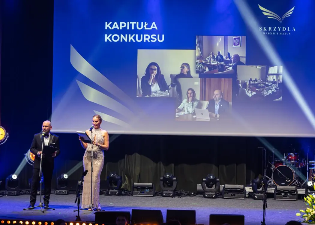 Gala Skrzydła Warmii i Mazur: prowadzący stoją na scenie, w tle zdjęcia z kapitułą konkursu