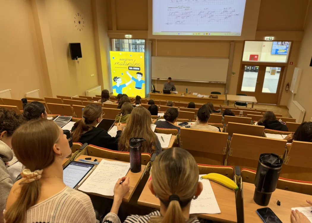 Sala wykładowa podczas warsztatów maturalnych na UWM; uczestnicy siedzą w ławkach i notują, a prowadzący pracuje przy tablicy i ekranie z zadaniami.