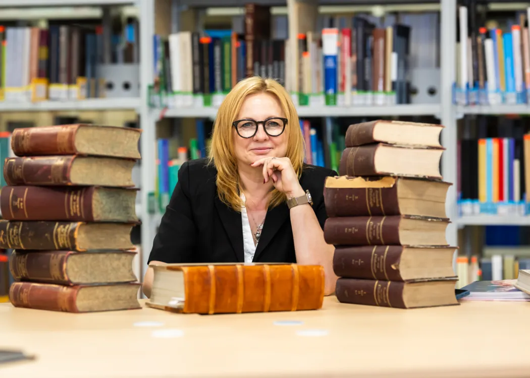 Kobieta siedzi przy biurku pełnym książek, za nią regały biblioteczne 