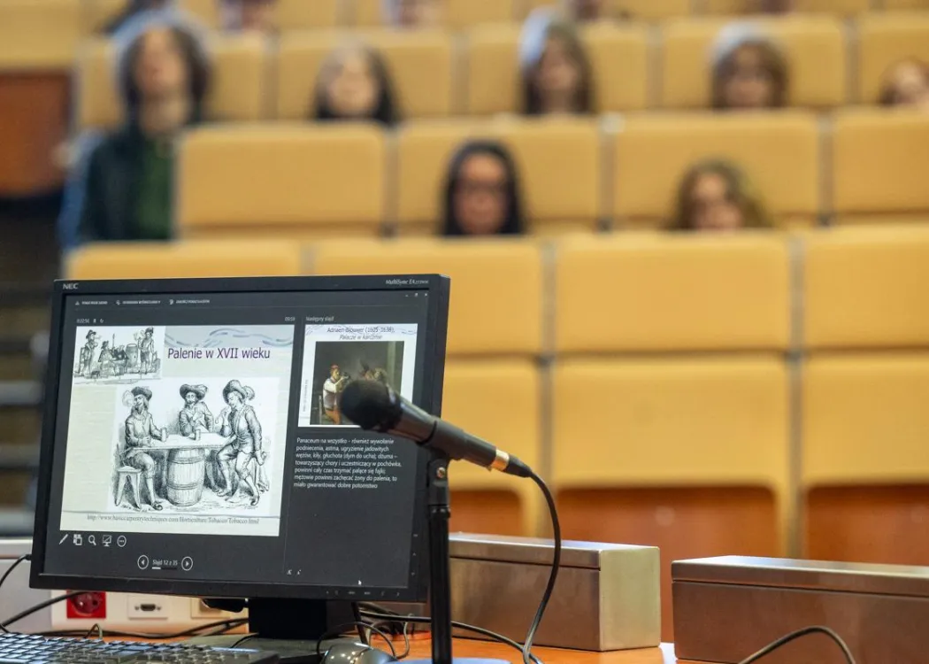 Zdjęcie przedstawia monitor komputera z prezentacją podczas wydarzenia "Czwartki z humanistyką", a w tle siedzą uczniowie szkół ponadpodstawowych biorący udział w wydarzeniu. 