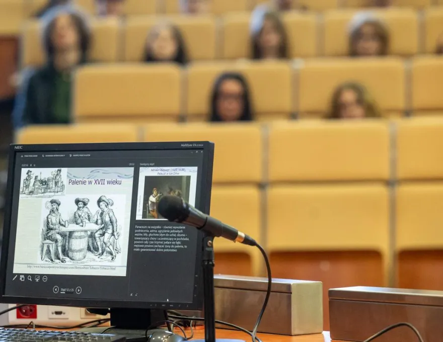 Zdjęcie przedstawia monitor komputera z prezentacją podczas wydarzenia "Czwartki z humanistyką", a w tle siedzą uczniowie szkół ponadpodstawowych biorący udział w wydarzeniu. 
