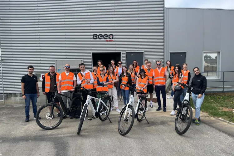 Studenci  stoją przed firmą produkujacą rowery, a rowery stoją przed nimi