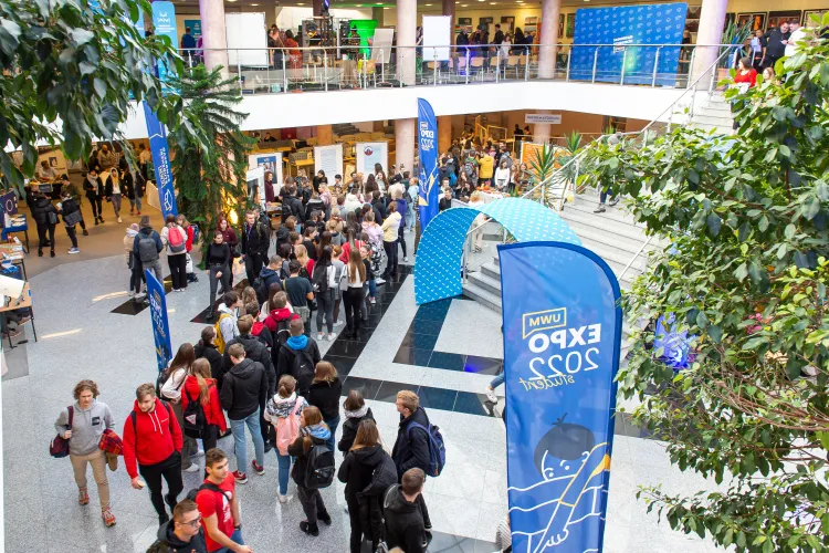 widok z góry na studentów stojących w bibliotece uniwersyteckiej podczas wydarzenia Student Expo, po prawej stoją niebieski flagi