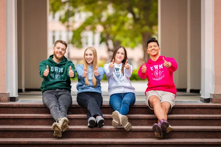 4 osoby w bluzach UWM, siedzą przed rektoracie z wystawionymi rękami, dają znak OK