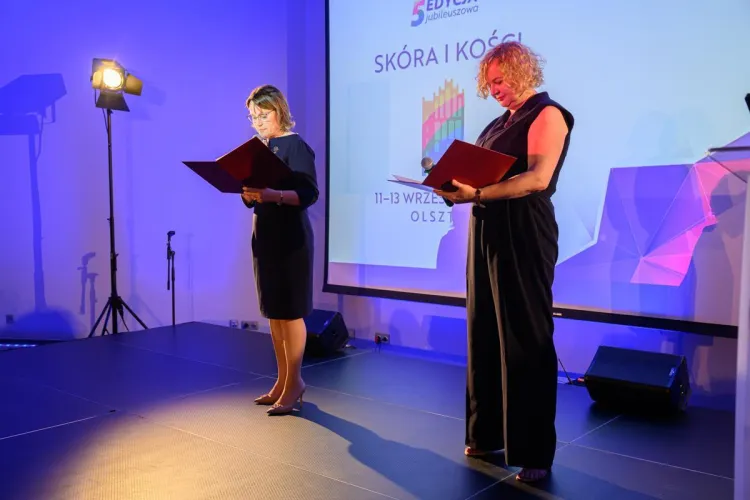 Na zdjęciu stoją dwie kobiety. Obie trzymają w rękach teczki. Pozują na tle ekranu z napisem "Skóra i kości"