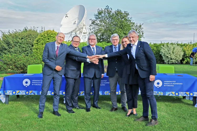 Sześcioro przedstawicieli polskiego konsorcjum projektu SKA Observatory stoi na tle anteny i trzyma dłonie położone jedna na drugiej