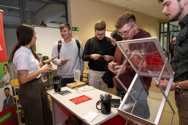 studenci stoją naprzeciwko stoiska i zbierają informacje o firmie