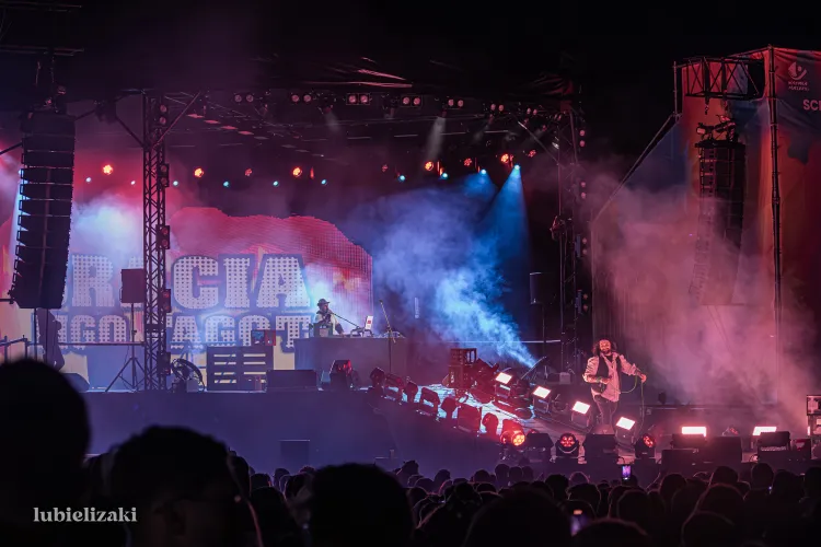 Scena z kolorowym oświetleniem na której występuje dwóch artystów - jeden na przodzie sceny śpiewa do mikrofonu, drugi z tyłu sceny gra na keyboardzie