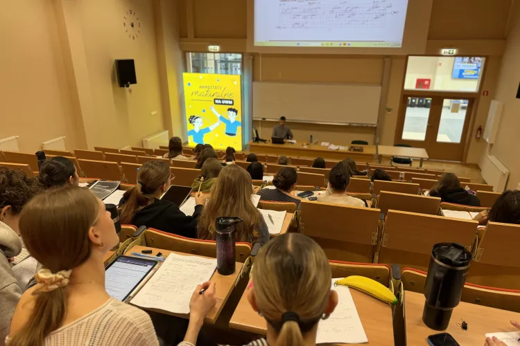 Sala wykładowa podczas warsztatów maturalnych na UWM; uczestnicy siedzą w ławkach i notują, a prowadzący pracuje przy tablicy i ekranie z zadaniami.