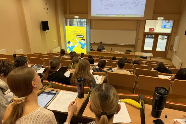 Sala wykładowa podczas warsztatów maturalnych na UWM; uczestnicy siedzą w ławkach i notują, a prowadzący pracuje przy tablicy i ekranie z zadaniami.