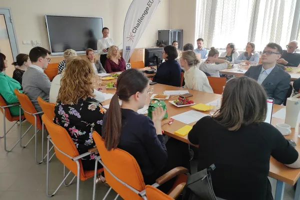 Grupa uczestników siedzących przy stole konferencyjnym podczas spotkania na Uniwersytecie Warmińsko-Mazurskim, słuchających prelegenta stojącego obok ekranu i baneru ChallengeEU.