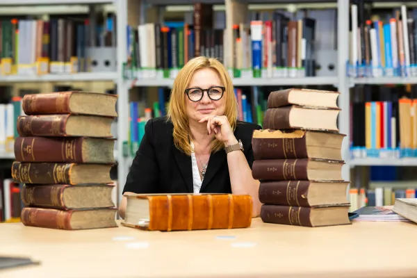 Kobieta siedzi przy biurku pełnym książek, za nią regały biblioteczne 