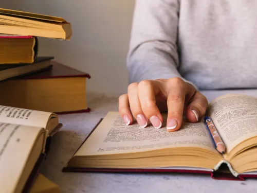 Osoba czyta książkę, wodząc palcem po stronie, obok leży stos książek