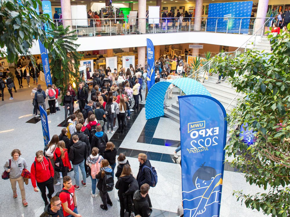 widok z góry na studentów stojących w bibliotece uniwersyteckiej podczas wydarzenia Student Expo, po prawej stoją niebieski flagi