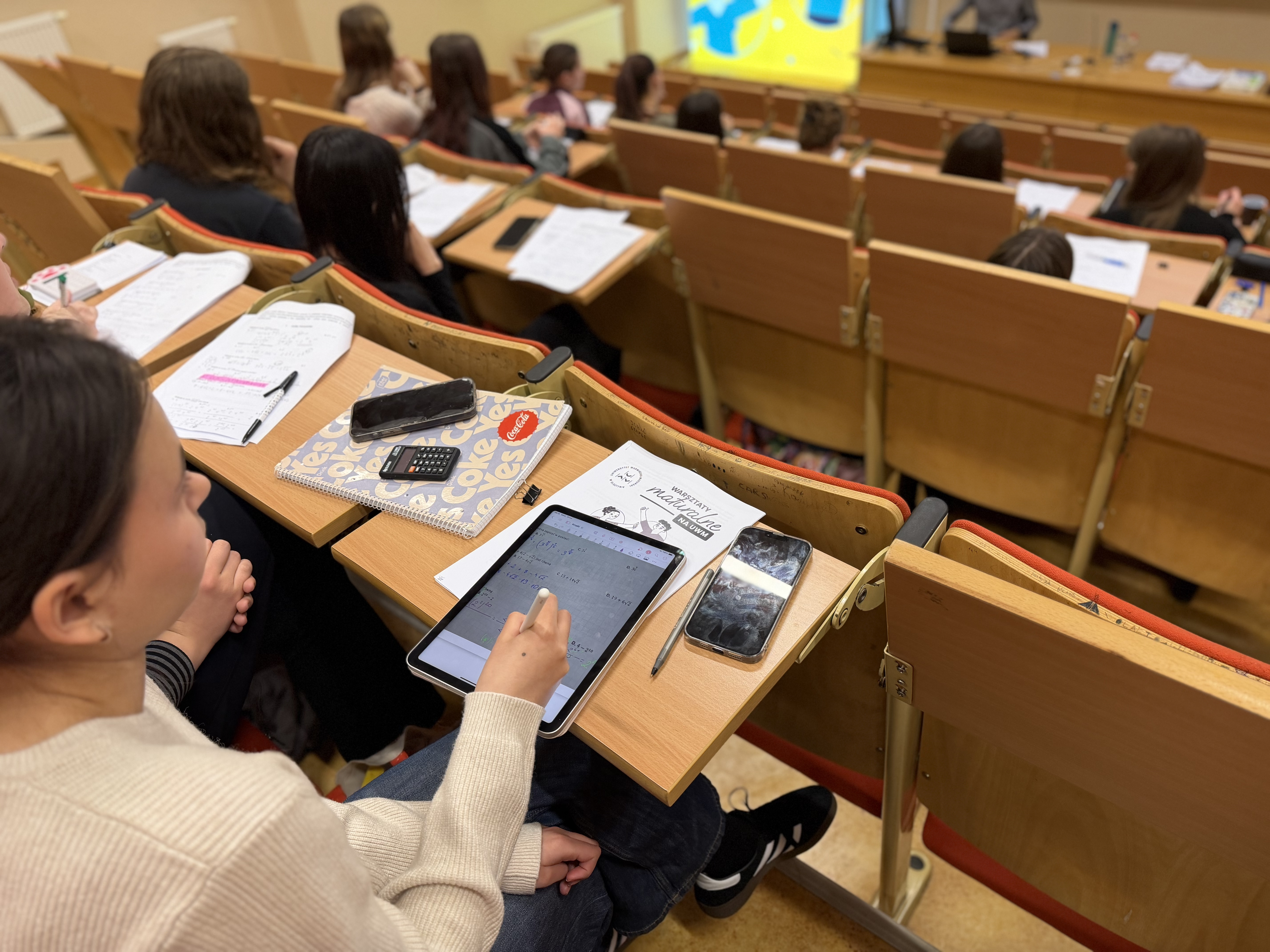 "Uczestnicy warsztatów siedzą w auli i rozwiązują zadania, korzystając z notatek, kalkulatorów i tabletu."