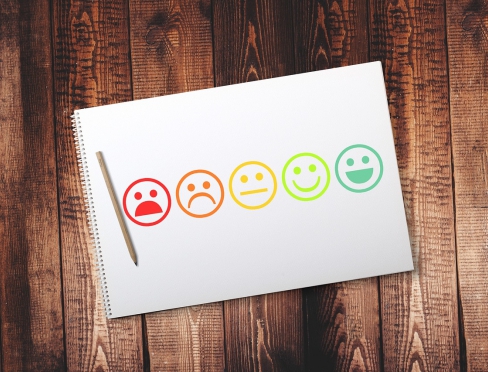 widziany z góry drewniany stół, a na nim kartka papieru z narysowanymi uśmiechniętymi i smutnymi buźkami oraz leżący obok długopis